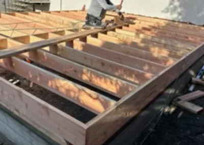 A construction worker measures and installs wooden beams on a new home's foundation framework on a sunny day.