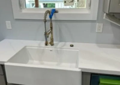 A modern farmhouse sink with a stylish gold faucet sits on a white countertop in a bright kitchen with a window view.