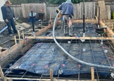 Two construction workers oversee pouring concrete into a rebar-framed foundation, surrounded by wooden forms and a black plastic cover.