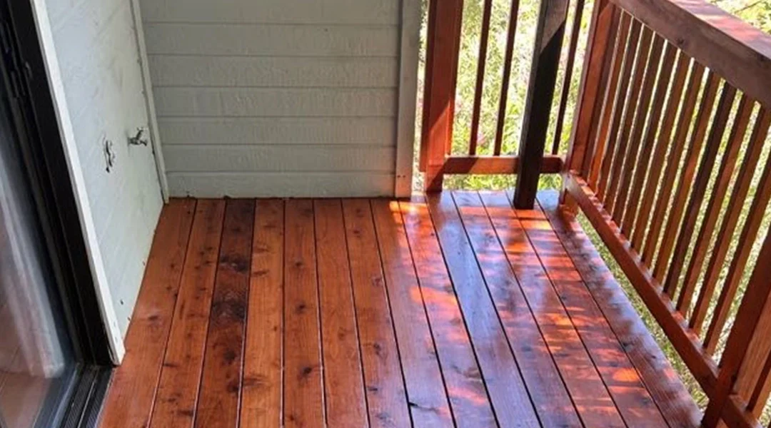 A wooden balcony with freshly stained planks, sunlight casting shadows on the smooth surface, and a railing in view.