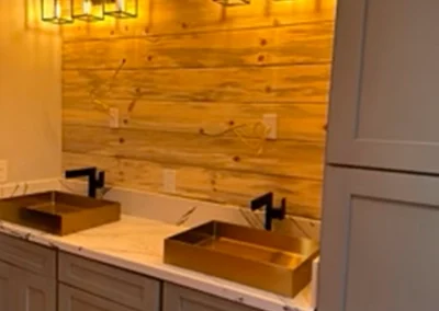 Modern bathroom with two gold-sink basins, marble countertop, and warm wood paneling illuminated by vintage-style light fixtures.