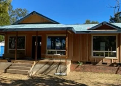 Front view of a rustic wooden house with a blue metal roof, large windows, and steps leading up, surrounded by trees under a clear blue sky.